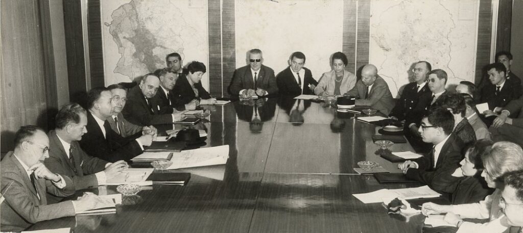 Photographie en noir et blanc datant de 1966. Dans une salle de réunion, une vingtaine de personnes sont réunies autour d'une table. Les visages sont concentrés.